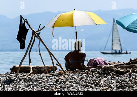 24 luglio 2019, Baden-Wuerttemberg, Gohren am Bodensee: una donna siede sulla riva del lago di Costanza sotto un ombrellone mentre un vele di imbarcazioni in background. Foto: Felix Kästle/dpa Foto Stock