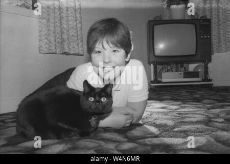 Anni '1980, storica, all'interno di una lounge, una giovane ragazza felice sdraiata sul tappeto con il suo gatto nero, Inghilterra, Regno Unito. All'angolo un televisore dell'epoca, con lettore VHS. Foto Stock