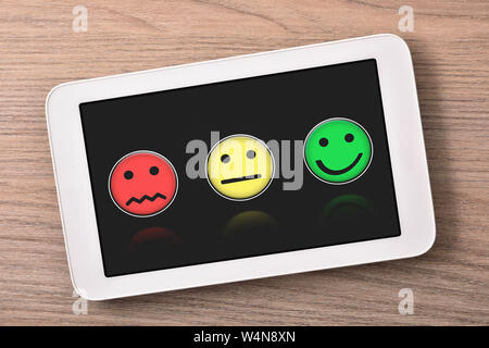 Soddisfazione dei livelli di rappresentazione con le emoticon sul tablet nero schermo sul tavolo di legno vicino. Composizione orizzontale. Vista dall'alto. Foto Stock