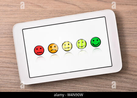 Soddisfazione dei livelli di rappresentazione con le emoticon su bianco tablet schermo sul tavolo di legno vicino. Composizione orizzontale. Vista dall'alto. Foto Stock