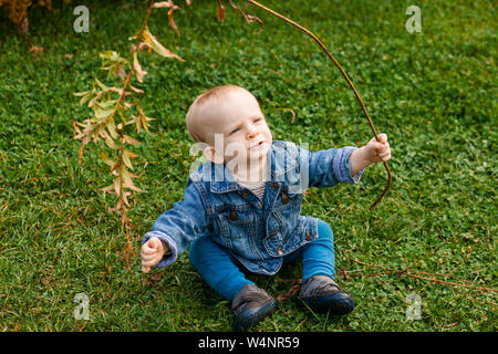 Un bambino si siede sul prato guardando verso l'alto e giocando con un ramo di albero Foto Stock