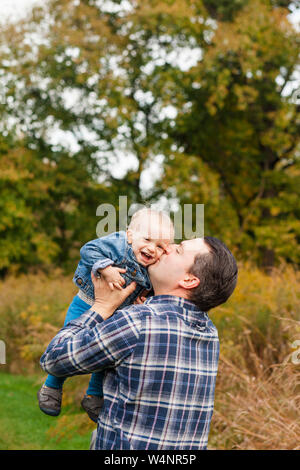 Un felice padre solleva il suo Laughing baby boy fino a per un bacio in un parco Foto Stock