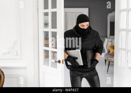 Ladro con nero balaclava rubare due moderni costosi computer portatili. Il ladro commette un reato in un appartamento di lusso con stucco. Foto Stock