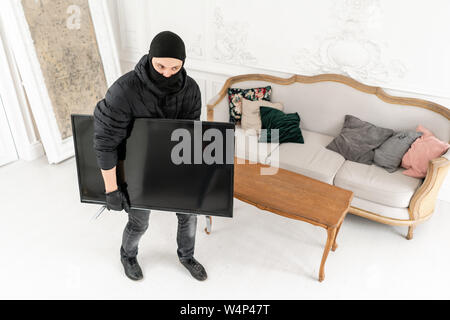 Il ladro commette un reato in un appartamento di lusso con stucco. Ladro con nero balaclava rubare moderno televisore costoso. Foto Stock