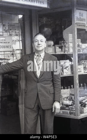 Degli anni Cinquanta, storico, un giornalaio maschio/toyshop proprietario in giacca e cravatta in piedi fuori l'ingresso il suo negozio, Inghilterra, Regno Unito. Foto Stock