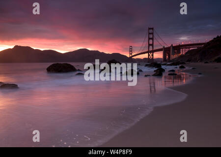 Di un bel colore rosa tramonto su una spiaggia con un ponte di sospensione della distanza Foto Stock