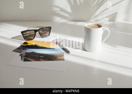 Tavolo bianco con penna, occhiali da sole, una tazza di caffè e le foto delle vacanze Foto Stock