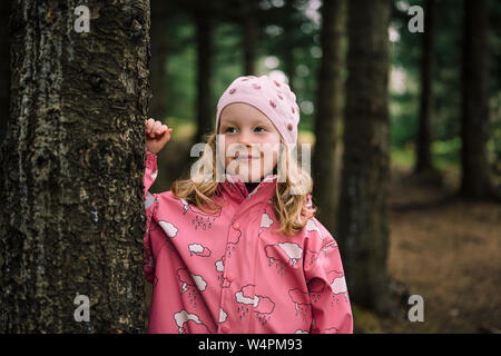 Bionda ragazza dai capelli rosa che indossa un impermeabile e cappello in piedi poggiando su albero a Reykjavik, Islanda foresta guardando lontano Foto Stock