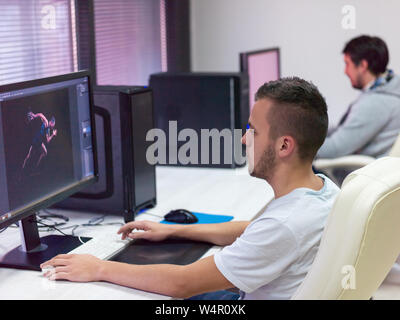 Giovani Graphic Designer che lavora al posto di lavoro. Ragazzo seduto a tavola il ritocco foto utilizzando tavoletta digitale con la penna e il computer desktop in ambienti chiusi. Freelan Foto Stock
