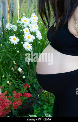 Baby bump profilo di una donna in stato di gravidanza tarda nel terzo trimestre in piedi nella parte anteriore del bianco e rosso dei fiori. Foto Stock
