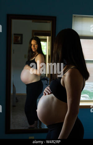 Donna incinta tardi nel terzo trimestre cercando longingly al suo bambino bump nello specchio. Ella è altamente anticipando l'arrivo del bambino. Foto Stock