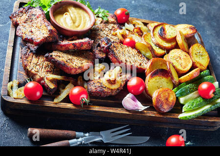 Close-up di Schwenkbraten, succosa, fumoso tedesco alla griglia costolette di maiale vengono cotti su una griglia oscillante servita su un rude tagliere con affettato pota fritto Foto Stock