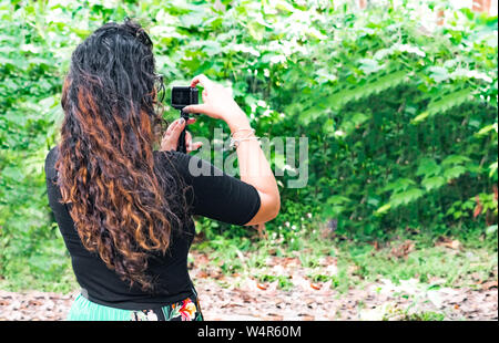 Trendy brunette botanico nel suo 20s con capelli ricci, il monitoraggio è la crescita di piante all'aperto durante il lavoro sul campo e facendo clic su foto dalla fotocamera di azione Foto Stock