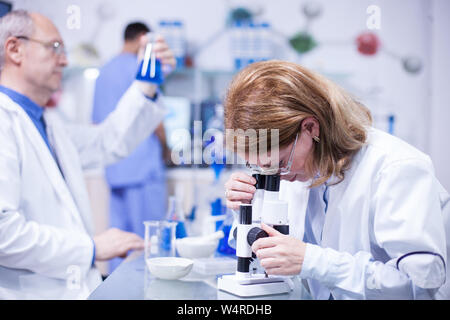 La mezza età femmina ingegnere chimico di lavorare su un microscopio in un laboratorio. Senior Scientist lavora con provette in background. Foto Stock