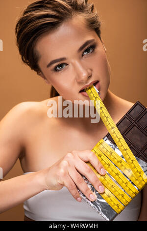 Giovane donna tenendo premuto barra di cioccolato e giallo nastro di misurazione in bocca isolato sul beige Foto Stock