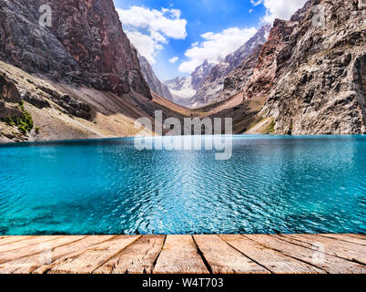 Tavolo in legno alto e lago di montagna con acqua turchese nella luce del sole sulla montagna rocciosa sfondo Foto Stock