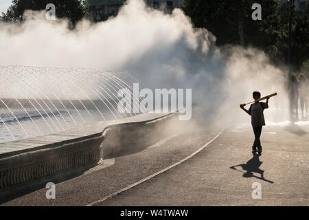 Piazza Unirii, Bucarest, Romania. Un bambino gioca con un acqua soaker (pompa) durante la torrida estate calore, davanti ad un'acqua di raffreddamento a spruzzo appannamento Foto Stock