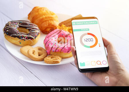 Calorie di conteggio e controllo degli alimenti concetto. donna utilizzando contacalorie applicazione sul suo smartphone con donut ,snack e cookie sulla piastra a bac Foto Stock