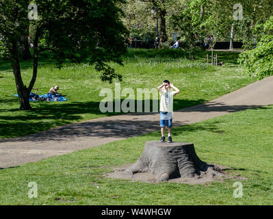 Un giovane ragazzo si erge su un ceppo di albero e guarda al cielo a St James Park, Londra centrale, in un giorno caldo e soleggiato. Foto Stock