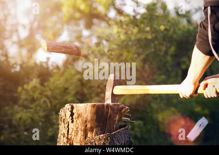 Un uomo in nero abbigliamento sportivo taglia una sharp ax log, alcune parti del quale sono sparse in microplacchette, illuminato dalla luce del sole nella foresta Foto Stock