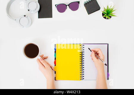 Donna workplace, mani femminili iscritto in un notepad, tenendo una tazza calda di caffè nero. Flatlay con cuffie, bicchieri, impianto ed un blocco note. Foto Stock
