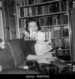 Giovane madre indossa un abito e blusa mantiene la sua figlia infante sul suo giro mentre è seduto su un divano nella libreria dei loro domestici, con una grande libreria in background, entrambi sorridenti, Detroit, Michigan, 1957. () Foto Stock