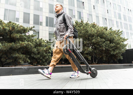 Attraente giovane uomo sorridente holding electric kick scooter mentre stava andando a fare una passeggiata lungo la strada. Foto Stock
