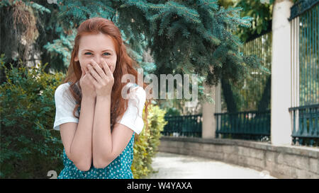 Lo zenzero Freckled ragazza sorride a uno scherzo. Sorridente bella donna redhead coperto la bocca con risate. Carino divertente i capelli rossi adolescente sorge nei pressi coniferou Foto Stock