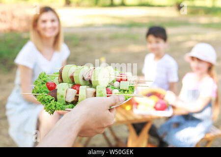 Uomo con gustosi piatti a base di carne e verdure sulla piastra per la sua famiglia all'aperto, primo piano Foto Stock