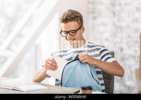 Studente maschio mettendo prenota in uno zaino a tavola Foto Stock