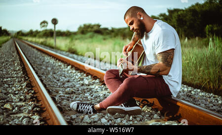 Giovane con tatuaggi e barba seduto su ferrovia e suonare la chitarra sul giorno nuvoloso Foto Stock