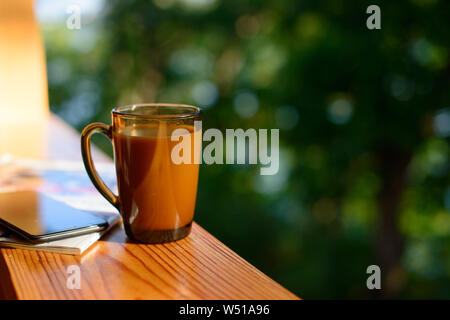 Tazza di caffè e cellulare sul davanzale più sfocata sullo sfondo di foresta al giorno d'estate e di sole. Copia gratuita dello spazio. Foto Stock