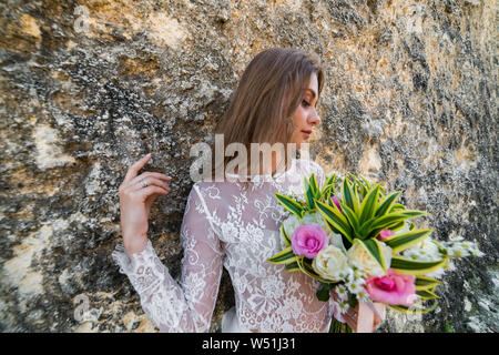 Bella Sposa in abito bianco azienda fiori e appoggiata contro la parete a strapiombo guardando al lato basso Foto Stock