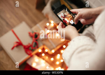 Mani tenendo il telefono e prendendo la foto del regalo di Natale scatole, santa hat, illuminazione luci su sfondo di legno nella camera oscura. Tanga elegante ragazza mi Foto Stock