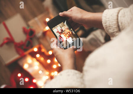 Mani tenendo il telefono e prendendo la foto del regalo di Natale scatole, santa hat, luci su sfondo di legno nella camera oscura. Tanga elegante ragazza in maglione mak Foto Stock