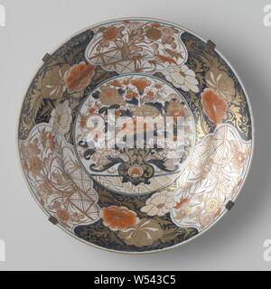 Piatto con un vaso di fiori e piante in fiore e cartigli floreali, piatto porcellana dipinta in underglaze blu e sullo smalto rosso e oro. Sul ripiano di un vaso di fiori con crisantemi e peonie, una stilizzata lotus sul vaso. La parete e edge sono suddivisi in sei scomparti alternativamente di piante fiorite dietro un recinto e peonie contro uno sfondo blu. La parete esterna con tre fiori spray. (Crisantemo, Prunus, peonia). Il fondo con proen. Il piatto è impostato in un telaio opaco. Imari., anonimo, Giappone, c. 1700 - c. 1750, Edo-periodo (1600-1868), porcellana (materiale), cobalto Foto Stock