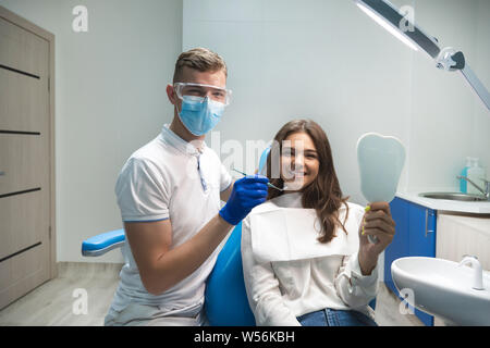 Bruna giovane donna paziente e dentista in blu guanti maschera medico e occhiali di sicurezza soddisfatto controllando i risultati del trattamento in clinica dentale Foto Stock