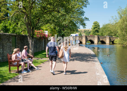 La gente che camminava lungo il lato delle rive Wye verso il ponte in una giornata di sole Bakewell Derbyshire England Regno Unito GB Europa Foto Stock