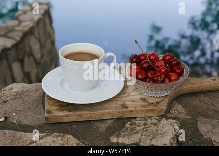 Le ciliegie e la tazza di caffè sul bordo di taglio sulla parete sopra il fiume Foto Stock