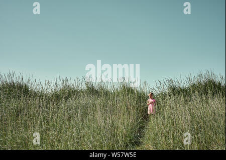 Campo il concetto di viaggio con graziosa bambina in abito rosa in piedi sul sentiero tra erba alta sulla grotta isola in estate Foto Stock