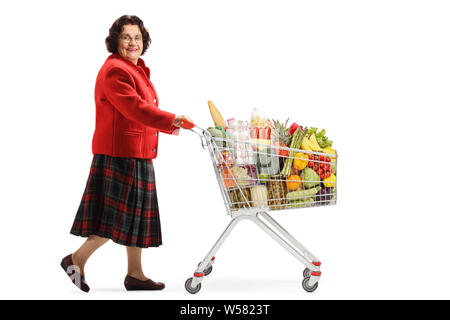 Lunghezza piena ripresa di profilo di un senior lady a piedi con un carrello della spesa con il cibo e sorridente alla telecamera isolata su sfondo bianco Foto Stock