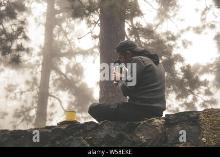 Giovani trekker prepara una bevanda calda facendo una sosta nella sua giornata esplorativa; sportivo da uomo seduto su una parete in un bosco nebbioso assaporare il profumo di caffè da Foto Stock