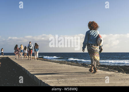 Gruppo di persone a piedi su un percorso di legno vicino al mare. I turisti torna a casa dopo una giornata in spiaggia. Gli amici sorseggiando una bella serata estiva togethe Foto Stock