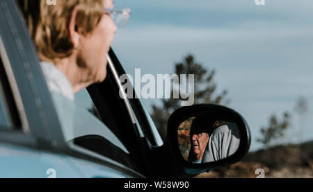 Una vecchia donna al di fuori della messa a fuoco con la sua testa fuori dal finestrino e concentrarsi sull'uomo anziano riflessa nella specchio auto godendo di una guida panoramica attraverso il mou Foto Stock