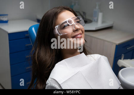 Sorridenti giovane donna paziente sdraiato sulla poltrona odontoiatrica indossando occhiali di sicurezza sotto la lampada medico in clinica Foto Stock