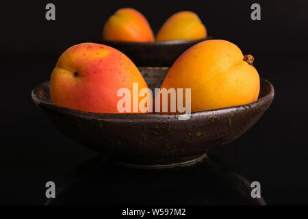 Il gruppo di quattro intere fresche Albicocche arancione in una buia la coppa in ceramica isolata sul vetro nero Foto Stock
