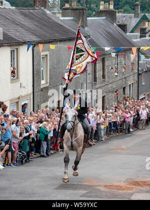 Langholm, Dumfries and Galloway, Scotland, Regno Unito. Il 26 luglio 2019. Il Corno, Henry Jeffrey, galoppa fino il Kirk Wynd portante il comune bandiera di equitazione. Foto Stock