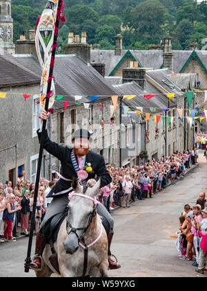 Langholm, Dumfries and Galloway, Scotland, Regno Unito. Il 26 luglio 2019. Il Corno, Henry Jeffrey, galoppa fino il Kirk Wynd portante il comune bandiera di equitazione. Foto Stock