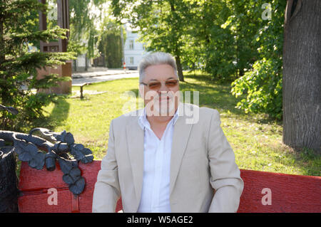 Ritratto di eldery uomo bello con i capelli grigi e gli occhiali seduto su un banco di lavoro in posizione di parcheggio Foto Stock