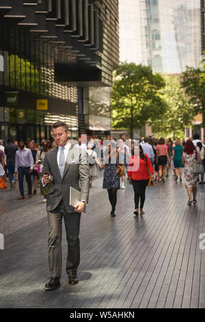 London, Regno Unito - Luglio, 2019. Un business man in tuta a piedi lungo uno di più di Londra, un complesso di uffici vicino al Municipio. Foto Stock